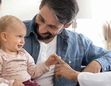 A baby smiling and sitting with her parents, alongside their pediatrician who is touching the baby's hand.