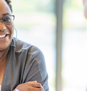 A middle-aged black woman smiling and leaning over a table, across from her female doctor.