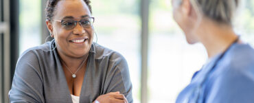 A middle-aged black woman smiling and leaning over a table, across from her female doctor.