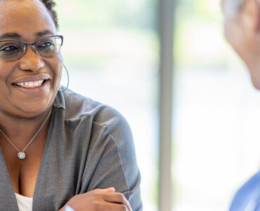 A middle-aged black woman smiling and leaning over a table, across from her female doctor.
