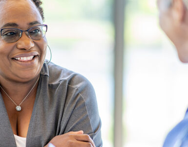 A middle-aged black woman smiling and leaning over a table, across from her female doctor.