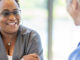 A middle-aged black woman smiling and leaning over a table, across from her female doctor.