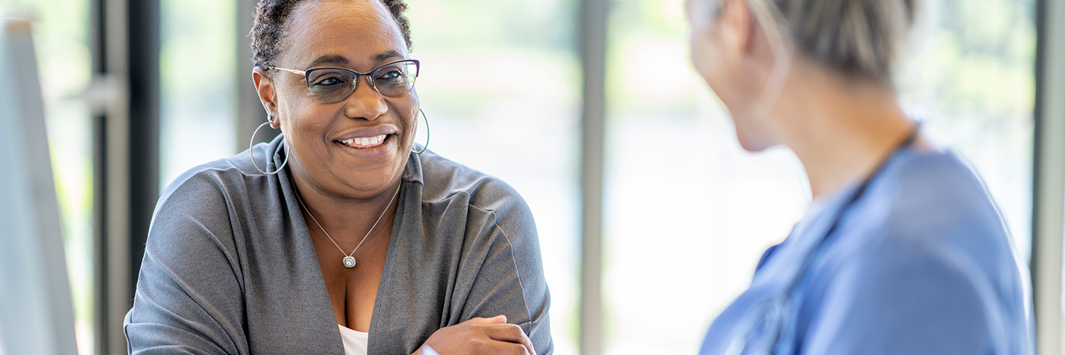 A middle-aged black woman smiling and leaning over a table, across from her female doctor.