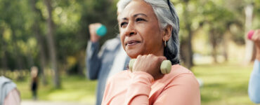 A middle-aged woman wearing a pink shirt, standing outdoors in an exercise glass and lifting a weight with one arm.
