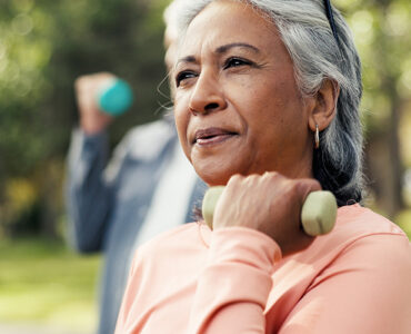 A middle-aged woman wearing a pink shirt, standing outdoors in an exercise glass and lifting a weight with one arm.