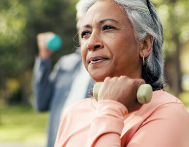 A middle-aged woman wearing a pink shirt, standing outdoors in an exercise glass and lifting a weight with one arm.