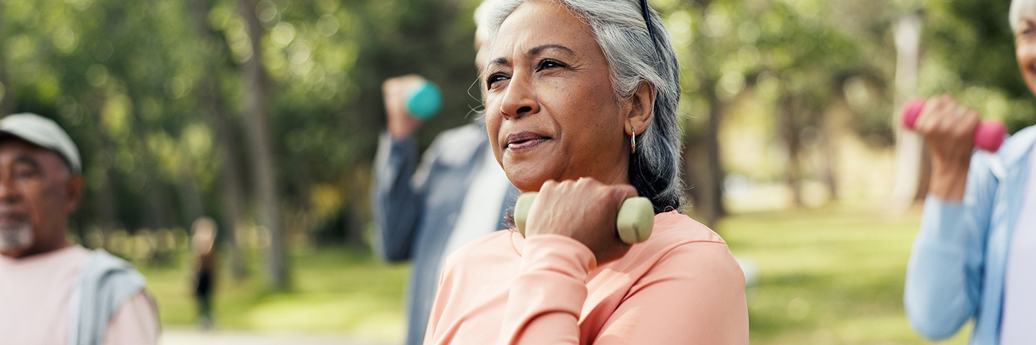 A middle-aged woman wearing a pink shirt, standing outdoors in an exercise glass and lifting a weight with one arm.