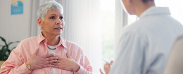 An older woman in her doctor's office, holding her heart while in a conversation.