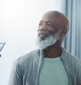 An older black man sitting in a medical office, smiling at the nurse who is holding a clipboard.
