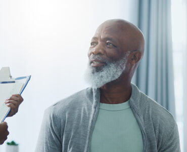 An older black man sitting in a medical office, smiling at the nurse who is holding a clipboard.
