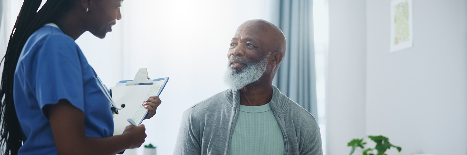 An older black man sitting in a medical office, smiling at the nurse who is holding a clipboard.