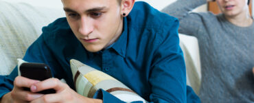 A teenage boy anxiously looking at his cell phone with his mother in the background looking concerned.