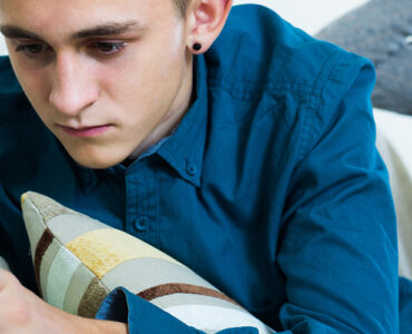 A teenage boy anxiously looking at his cell phone with his mother in the background looking concerned.