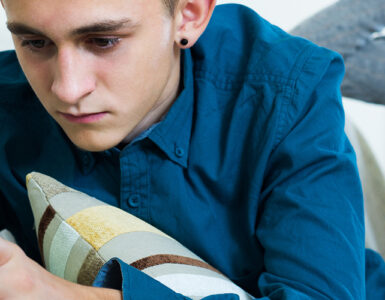 A teenage boy anxiously looking at his cell phone with his mother in the background looking concerned.