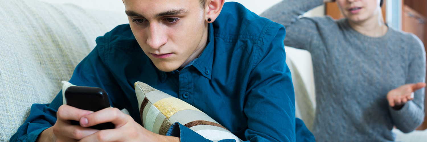 A teenage boy anxiously looking at his cell phone with his mother in the background looking concerned.