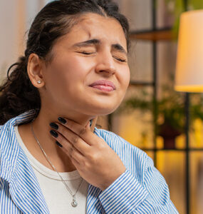 A woman holding her throat in pain while sitting at her desk at home.