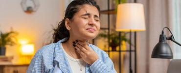 A woman holding her throat in pain while sitting at her desk at home.