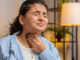 A woman holding her throat in pain while sitting at her desk at home.