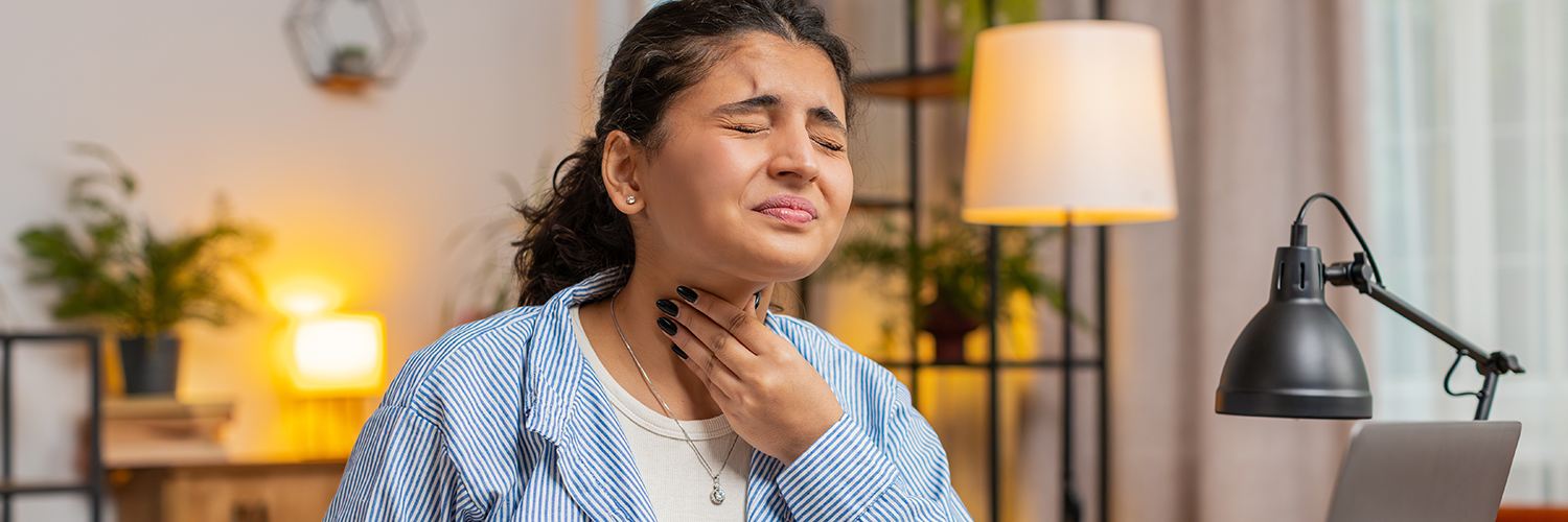 A woman holding her throat in pain while sitting at her desk at home.