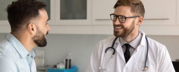 A male patient in his 40s speaking to a male doctor in his medical office, both smiling.