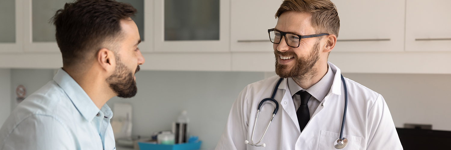 A male patient in his 40s speaking to a male doctor in his medical office, both smiling.