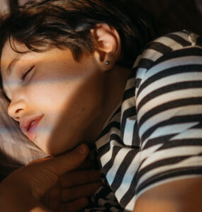 A teenage boy wearing a striped shirt, sleeping in his bed.