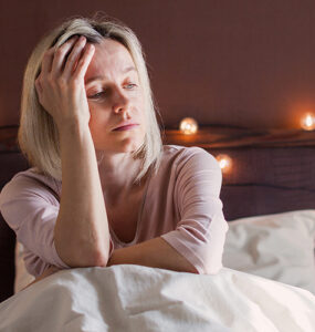 A woman in her 40s sitting in her bed, holding her head and looking tired.
