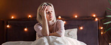 A woman in her 40s sitting in her bed, holding her head and looking tired.
