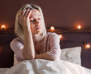 A woman in her 40s sitting in her bed, holding her head and looking tired.