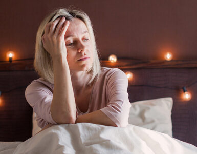 A woman in her 40s sitting in her bed, holding her head and looking tired.