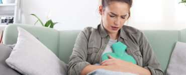 A young woman sitting on her couch with a heating pad pressed against her stomach.