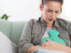 A young woman sitting on her couch with a heating pad pressed against her stomach.