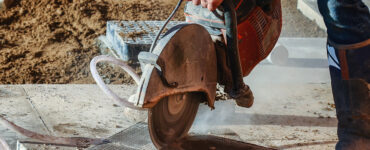 Construction worker using a circular saw to cut concrete, producing dust during outdoor construction work.