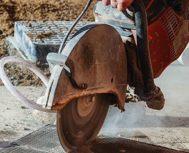 Construction worker using a circular saw to cut concrete, producing dust during outdoor construction work.