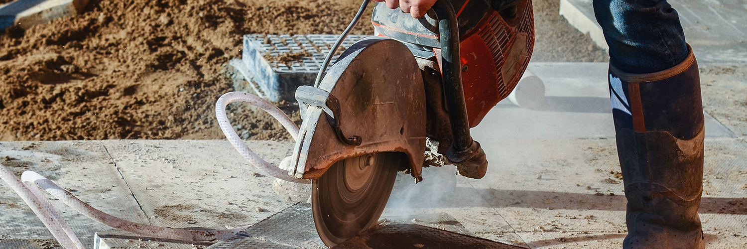 Construction worker using a circular saw to cut concrete, producing dust during outdoor construction work.