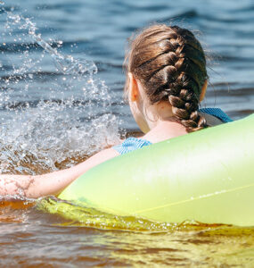 A young girl swimming in the bay within a yellow tube, with her back and braided hair in view.