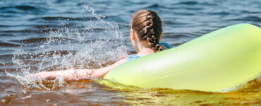 A young girl swimming in the bay within a yellow tube, with her back and braided hair in view.