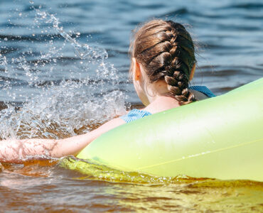 A young girl swimming in the bay within a yellow tube, with her back and braided hair in view.