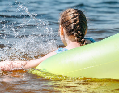 A young girl swimming in the bay within a yellow tube, with her back and braided hair in view.