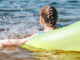 A young girl swimming in the bay within a yellow tube, with her back and braided hair in view.