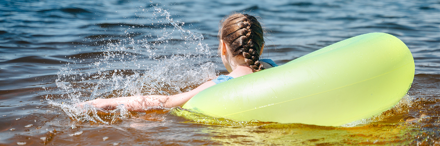 A young girl swimming in the bay within a yellow tube, with her back and braided hair in view.