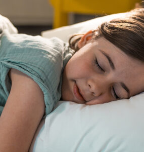 A young girl sleeping in her bed, wearing a green shirt with white bed sheets.