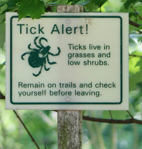 A sign outside in a wooded area warning hikers that ticks are present, with "Tick Alert!"