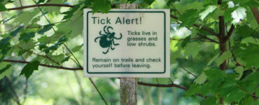 A sign outside in a wooded area warning hikers that ticks are present, with "Tick Alert!"