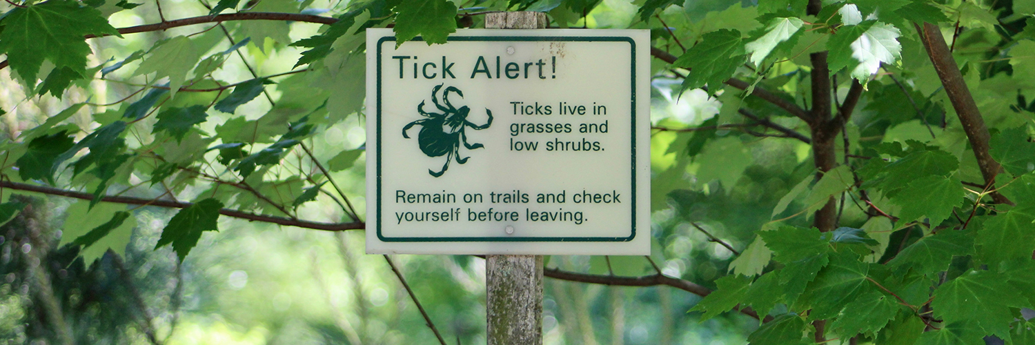 A sign outside in a wooded area warning hikers that ticks are present, with "Tick Alert!"