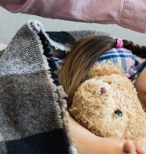 A young child laying in bed, holding her stuffie while her mom rubs her head.