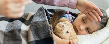 A young child laying in bed, holding her stuffie while her mom rubs her head.