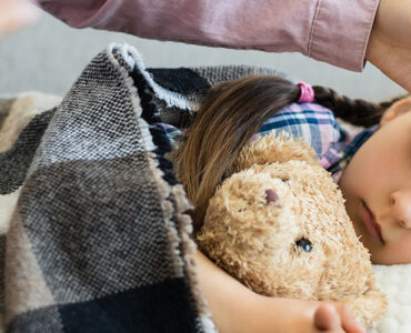 A young child laying in bed, holding her stuffie while her mom rubs her head.