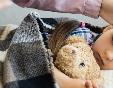 A young child laying in bed, holding her stuffie while her mom rubs her head.