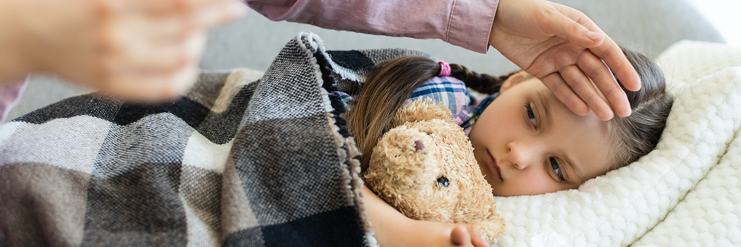 A young child laying in bed, holding her stuffie while her mom rubs her head.